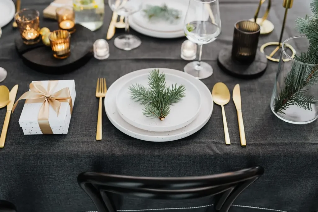 Dark charcoal linen Christmas table with white plates, a pine sprig, gold cutlery and amber candle holders