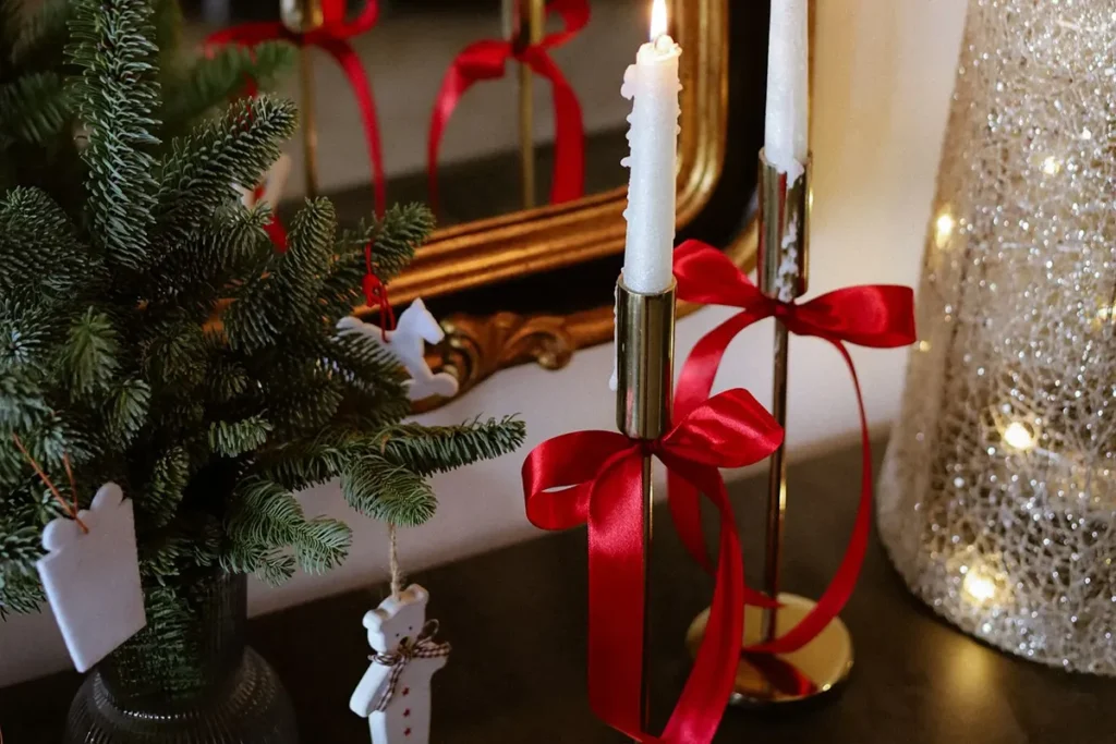 White taper candles with red ribbon bows in brass holders beside a gold mirror and a fir sprig