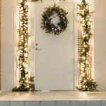 Christmas front porch with a wreath on a white door, lit garland frame, two potted trees and warm string lights