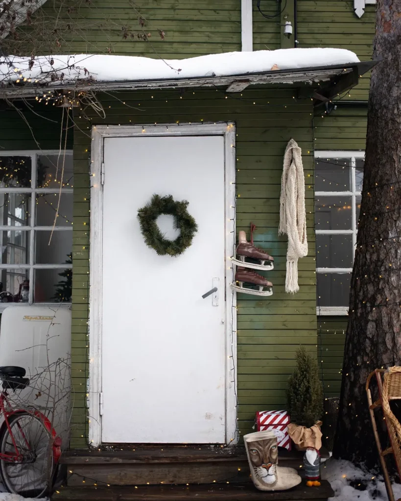 White front door with a simple wreath, string lights and a small spruce on a snowy wooden porch