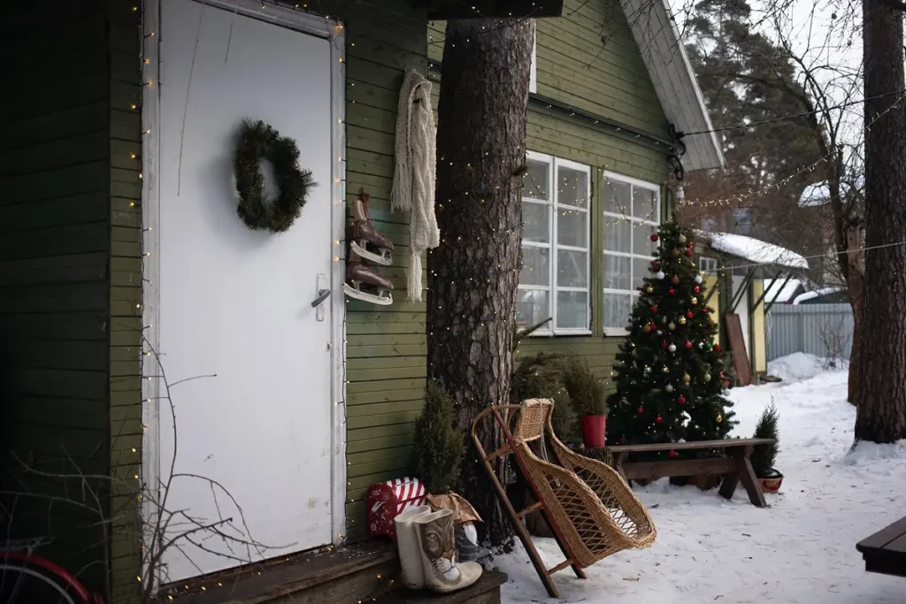 Christmas porch with a wicker sled, a wooden bench, a small tree and string lights beside a white door in the snow