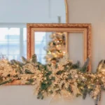 Christmas mantel decor with a full garland, warm white lights, taper candles and an arched mirror above a floating mantel shelf