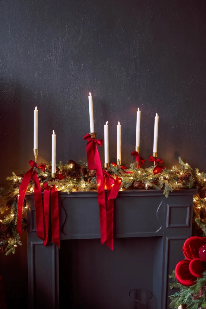 Dark Christmas mantel with a lit garland and white taper candles tied with red ribbon bows