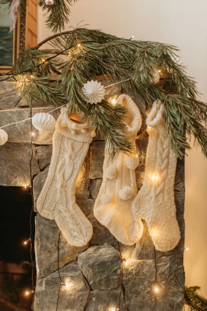 Three matching cream knit stockings hung under a pine garland with warm lights on a stone fireplace
